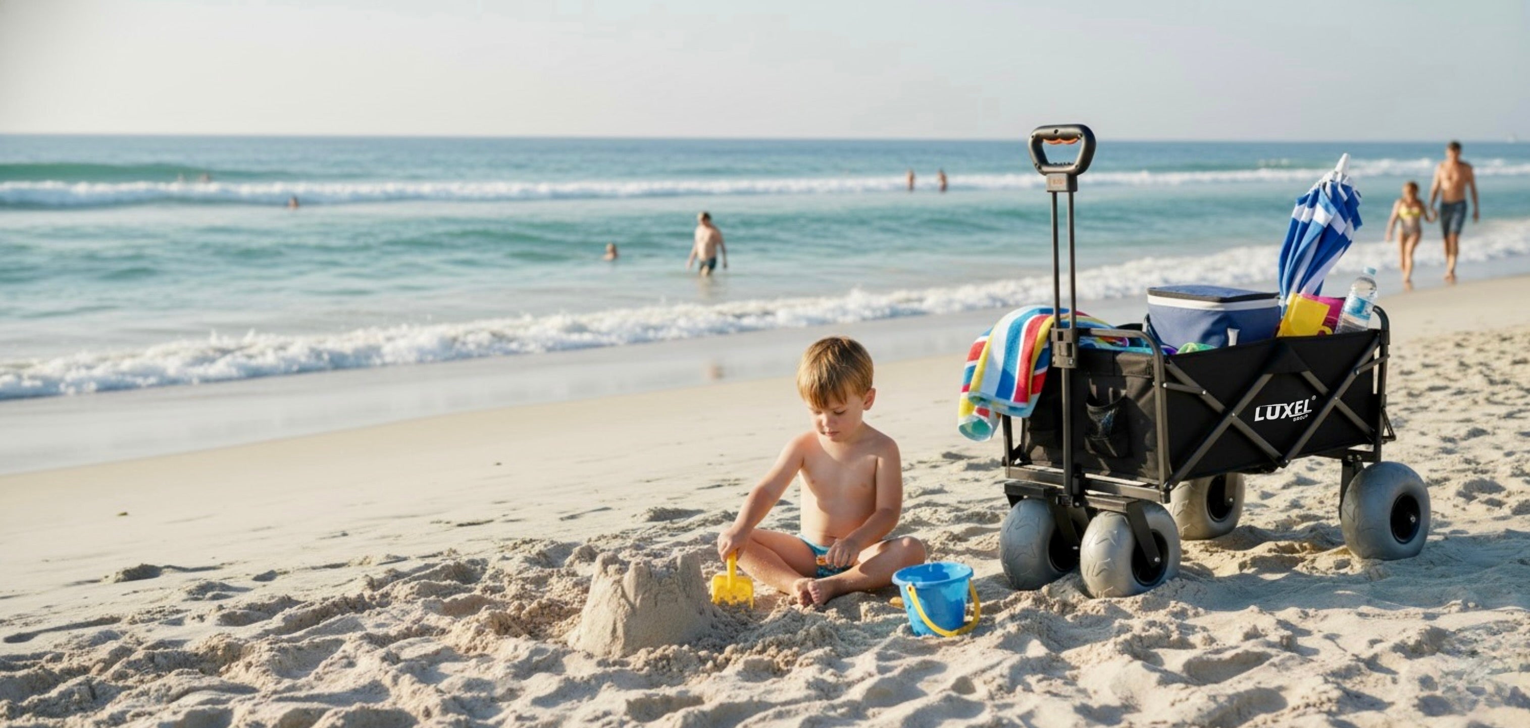 Child playing on a sandy beach with a beach cart and sand toys, ocean in the background.