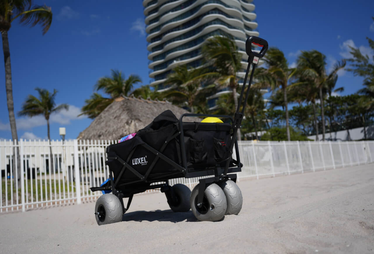 Luxel Group electric beach wagon with gray balloon wheels and loaded cargo, standing on a sandy beach with palm trees and modern building in background.