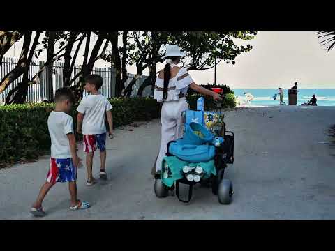 Woman in white outfit and hat walking towards the beach with two children, pulling a fully loaded Luxel Group electric wagon with large gray sand wheels carrying towels, beach gear, and bags.
